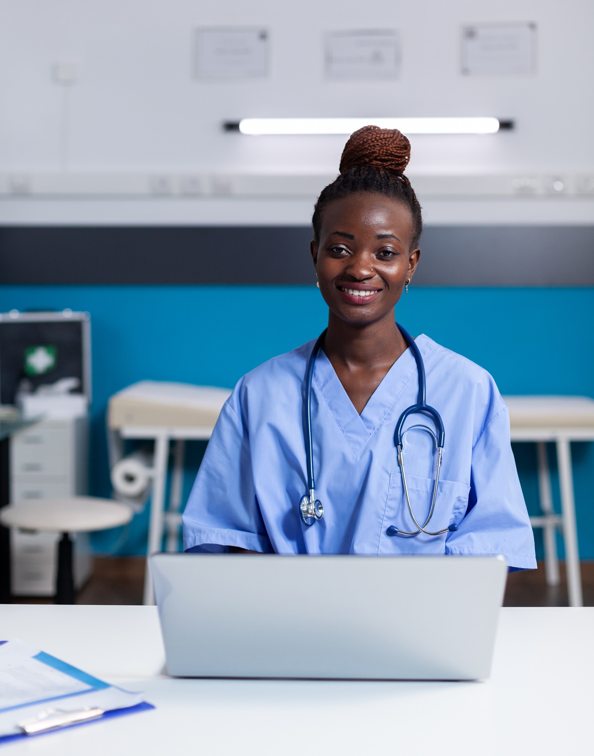 Portrait of african nurse using laptop at white desk sitting in medical office. Black woman with assistant occupation wearing uniform and working at healthcare clinic