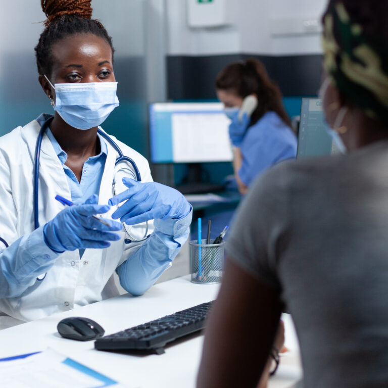 African therapist doctor discussing with sick patient during clinical consultation explaining medical signing paperwork in hospital office. Women wearing protective face mask against covid19